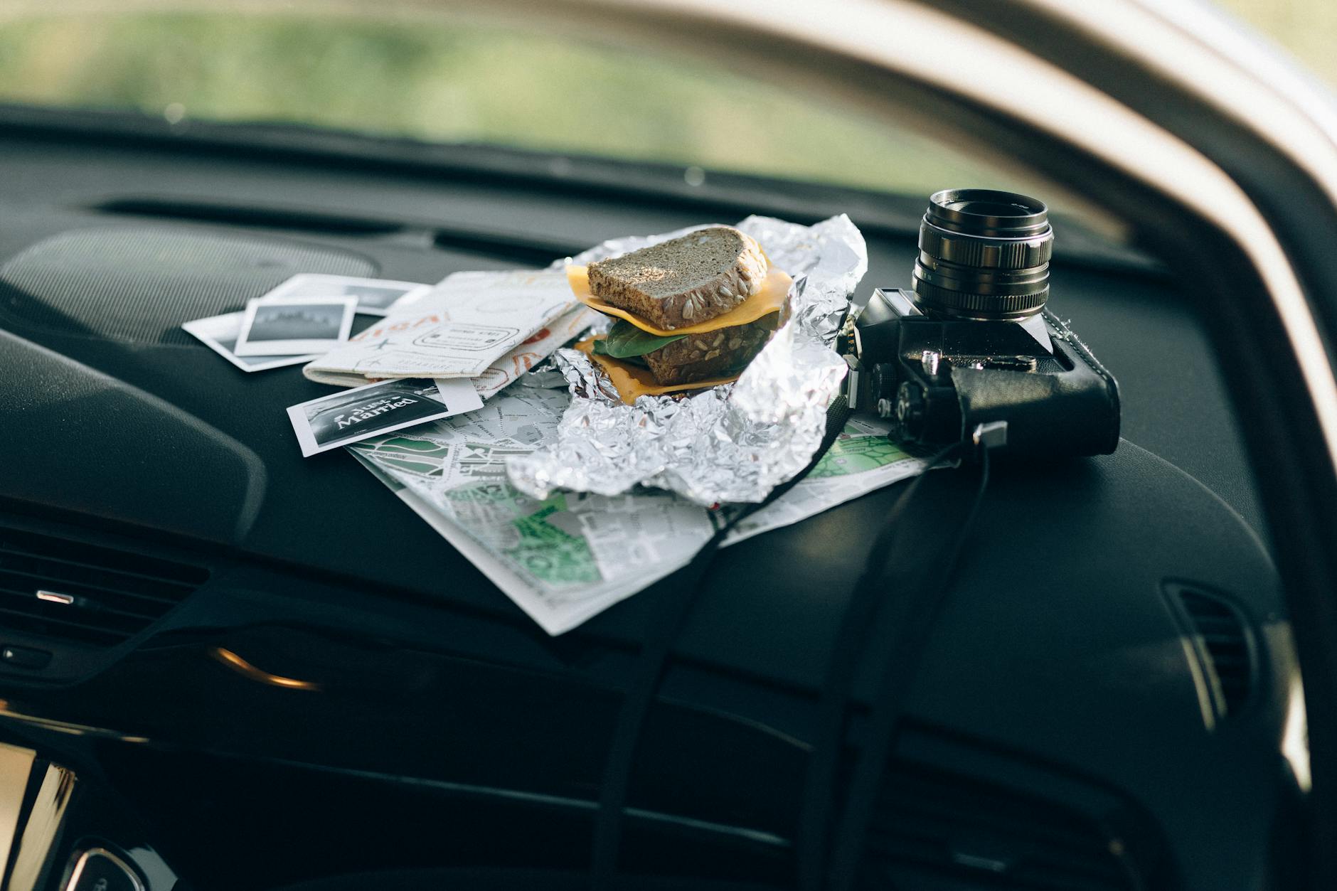 A camera and a sandwich wrapped in foil placed on a car dashboard with maps and photos.