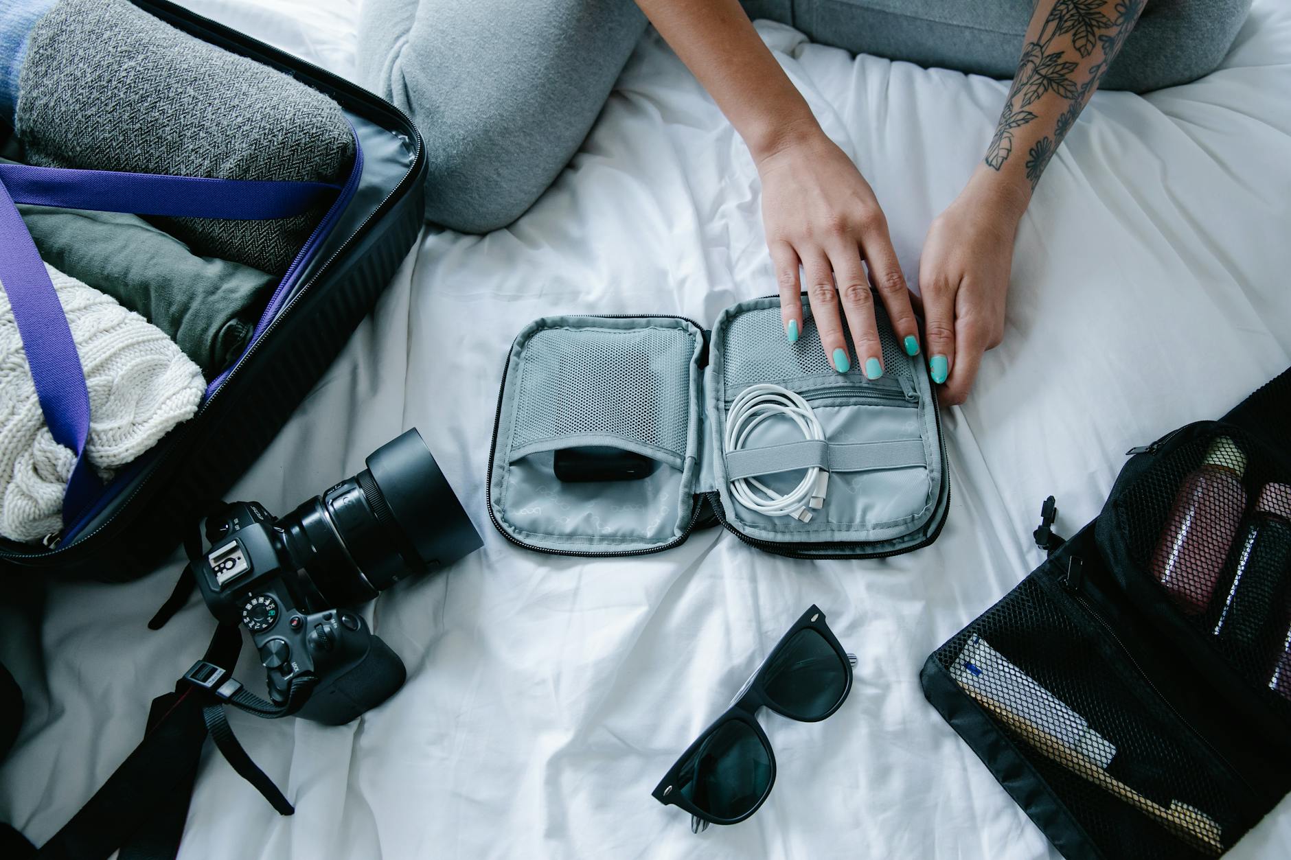 Woman with tattoos packs electronics, camera, and sunglasses in a suitcase on a bed.